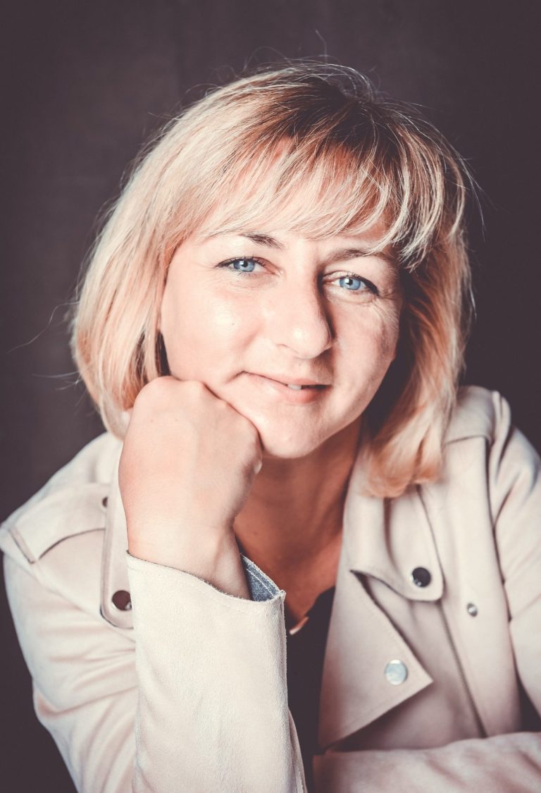 Porträt einer blonden Frau mit blauen Augen in heller Bikerjacke, die lächelnd ihr Kinn auf die Hand stützt.