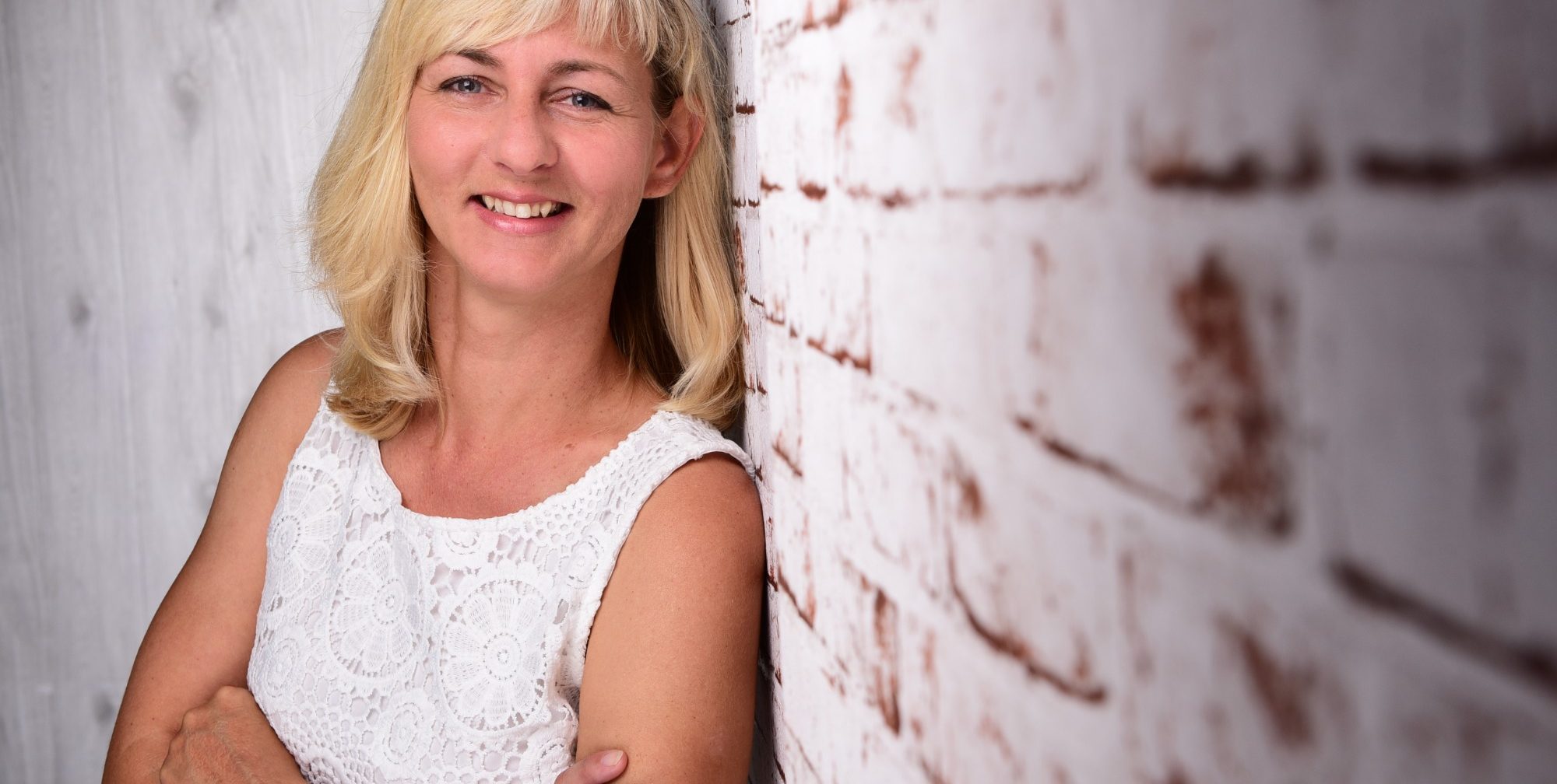 Porträt einer lächelnden Frau mit blonden Haaren. Sie trägt ein weißes Oberteil und lehnt seitlich an einer hellen Backsteinwand.
