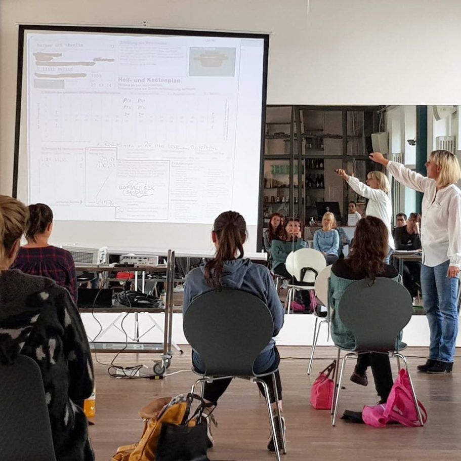 Eine Referentin steht vor einer Leinwand und erklärt einer Gruppe von Teilnehmenden einen projizierten Heil- und Kostenplan. 